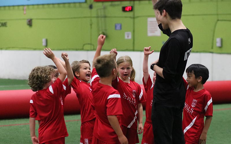 A group of kids in Skills Institute soccer uniforms do a group cheer with their instructor