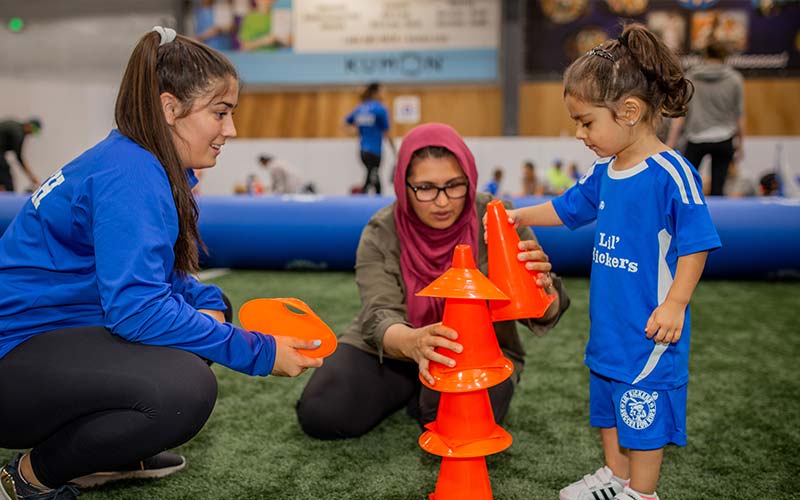 A young child practices hand-eye coordination with soccer cones in a Lil' Kickers soccer class while a parent watches