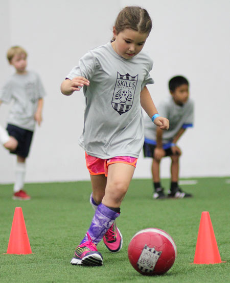 A camper navigates kicking a soccer ball through orange cones in a Skills Institute drill